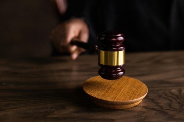 A close-up of a brown wooden gavel resting on a matching wooden table. The gavel is positioned with its head facing the viewer, symbolizing justice and legal authority.