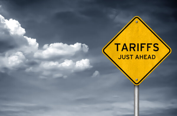 Impact road sign that says “TARIFFS JUST AHEAD” with cloudy skies in the background.